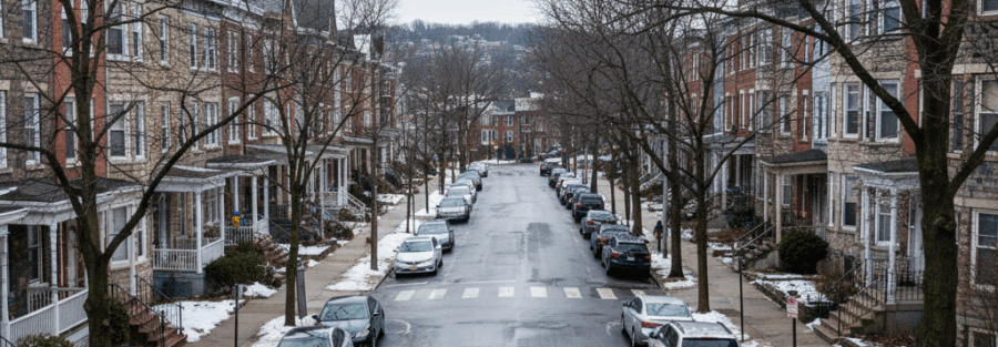 Cars driving and parked along a hilly residential street in Roxborough/Manayunk with Philadelphia-style rowhomes in light winter conditions.